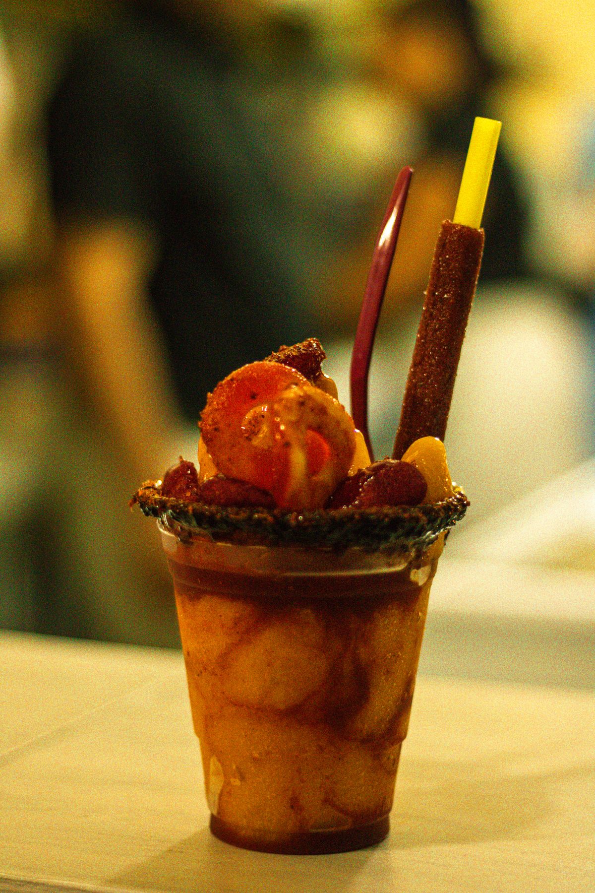 A cup of fruit drenched in chamoy with a chilli-dusted tamarind stick standing in it.